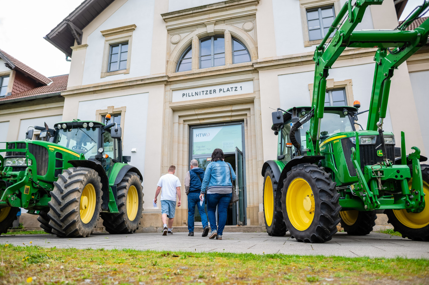 Zwei Traktoren vor einem Gebäude, zwischen den Traktoren laufen 3 Personen auf den Eingang des Gebäudes zu.