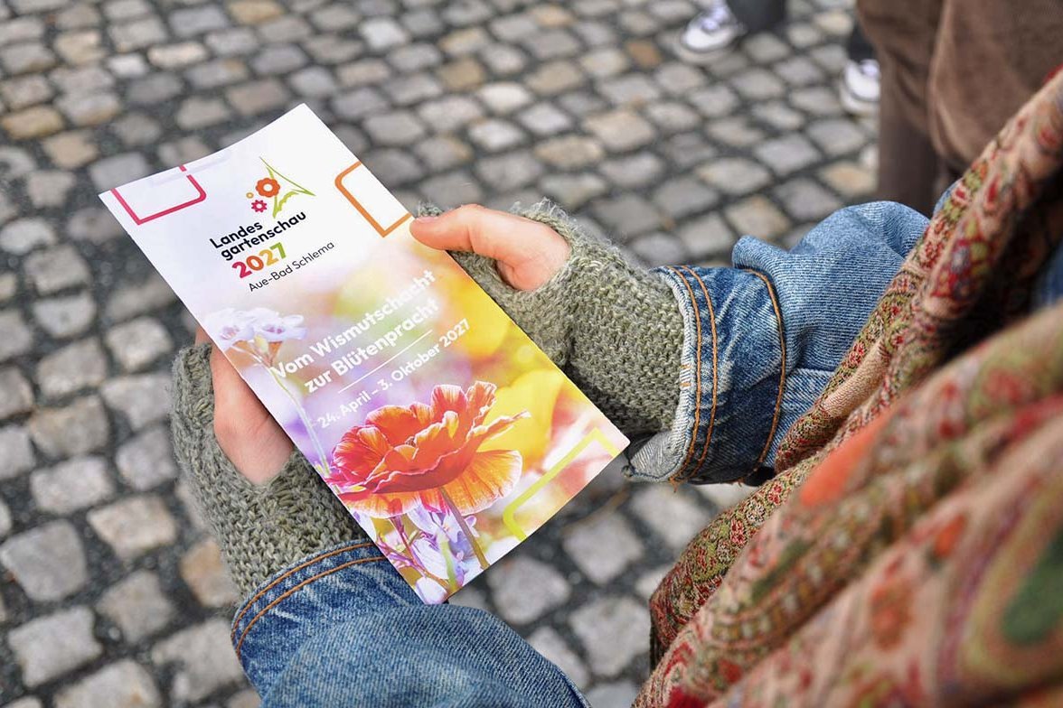 ): Eine Studierende hält den Flyer der Landesgartenschau 2027 in der Hand. Darauf ist das Motto „Vom Wismutschacht zur Blütenpracht“ zu lesen