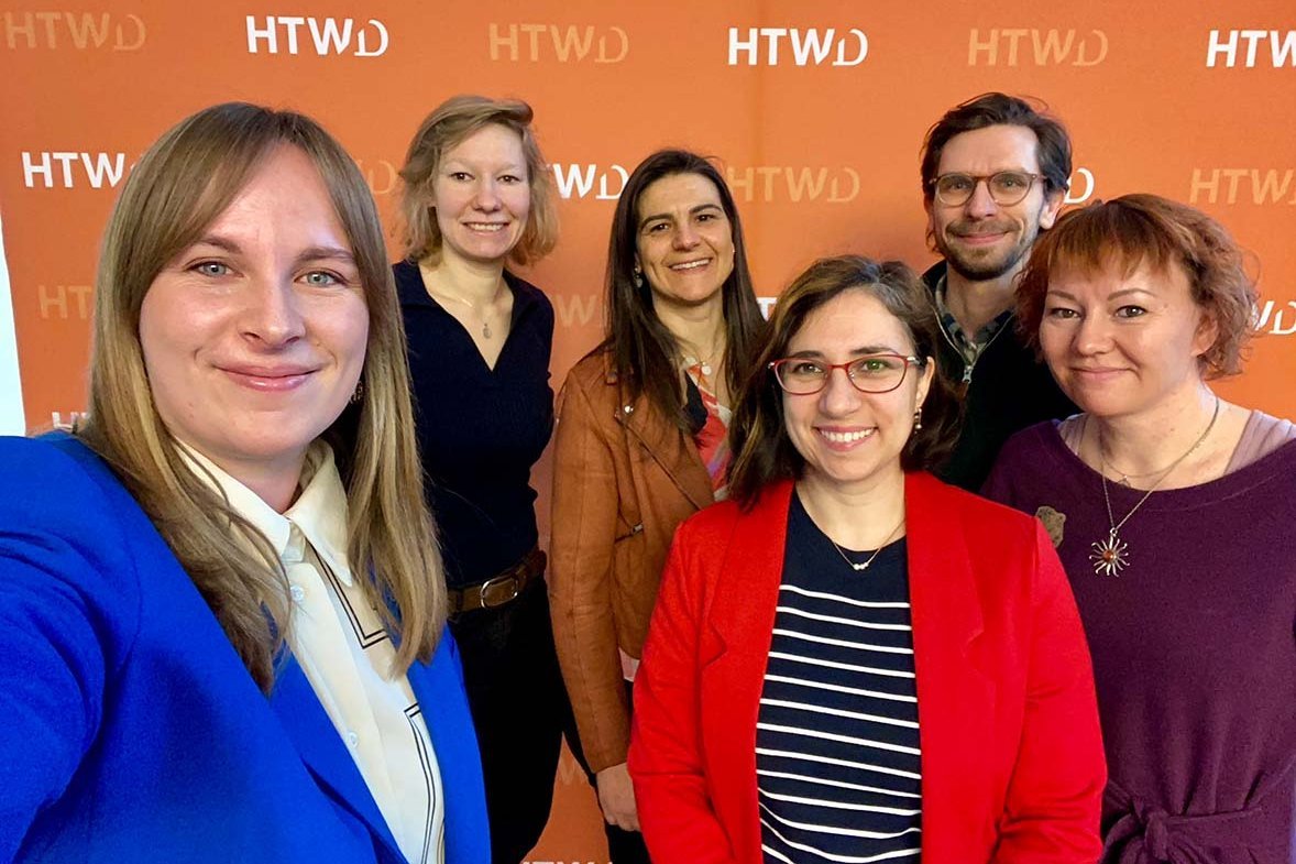 Fünf weiblich gelesene Personen und eine männlich gelesene Person stehen vor einer HTWD-Pessewand und lächeln den